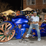 Custom blue bagger motorcycle with oversized bronze front wheel parked at Black Bike Week in Daytona Beach while a rider sits on the bike surrounded by vendors and crowds at night.