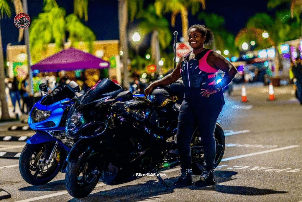 Shena Carty, motorcycle rider known as Showstopper, standing beside a black Suzuki Hayabusa at a bike night with other motorcycles in the background.